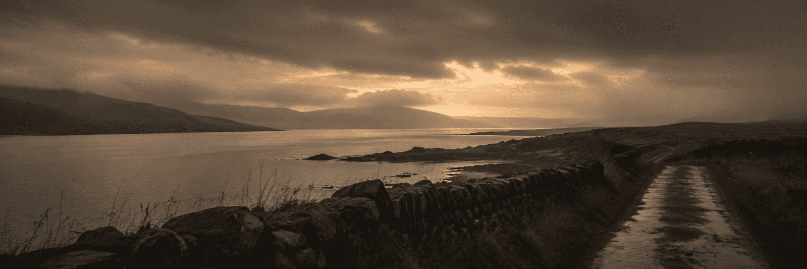 A narrow wet coastal road runs beside a stone wall, overlooking a rocky shoreline and calm sea beneath dark storm clouds with sunlight breaking through in the distance.